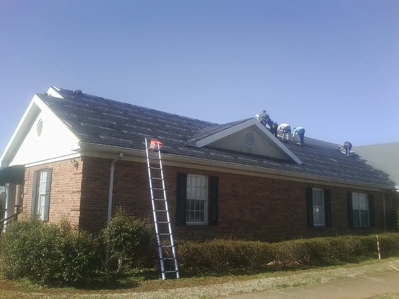 Crew tearing shingles off a brick ranch roof with a ladder set — Metal Roof Repair work by our crew in Speonk