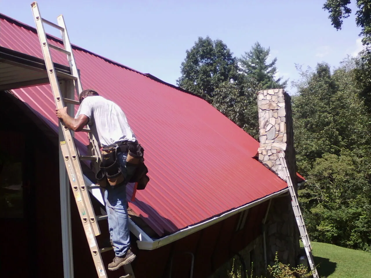 Roofer on a ladder beside a red metal roof and stone chimney — Soffit & Fascia Repair by Roofing Brothers in Speonk