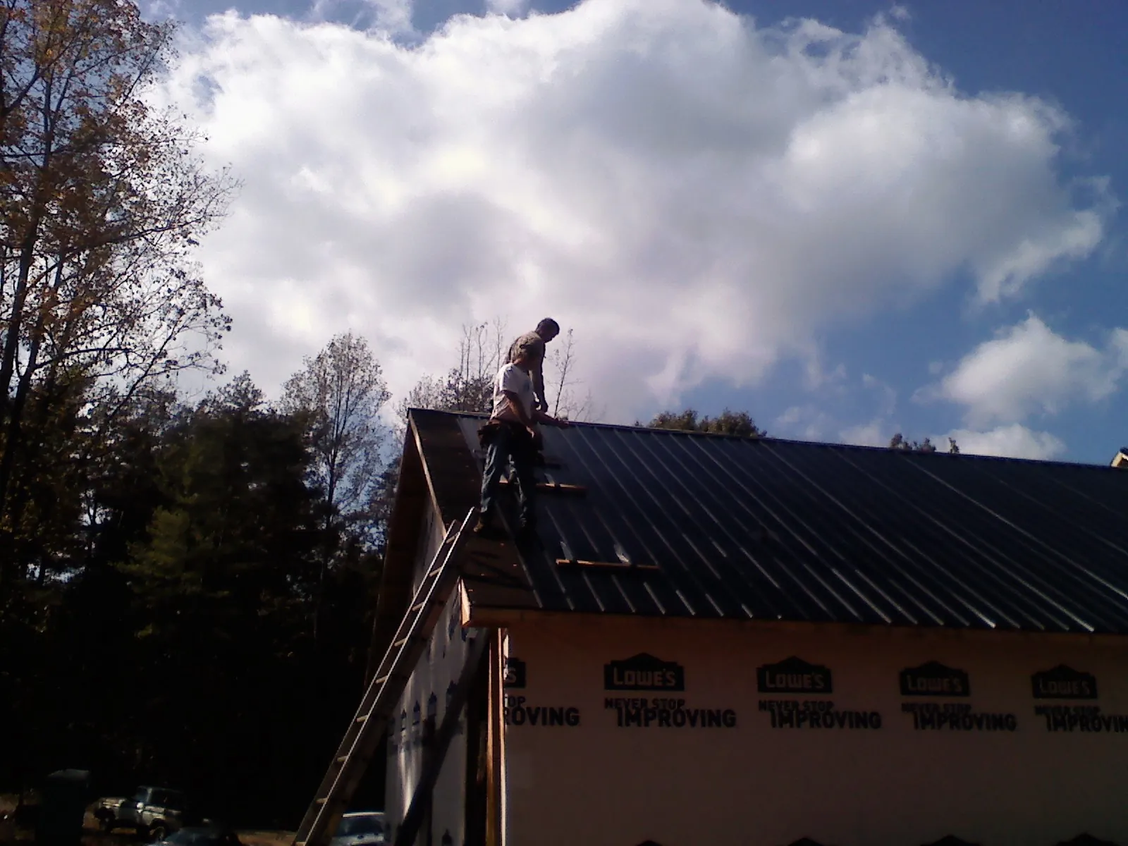 Roofer fastening dark metal panels high against a cloudy sky, from a Roof Inspection job in Speonk